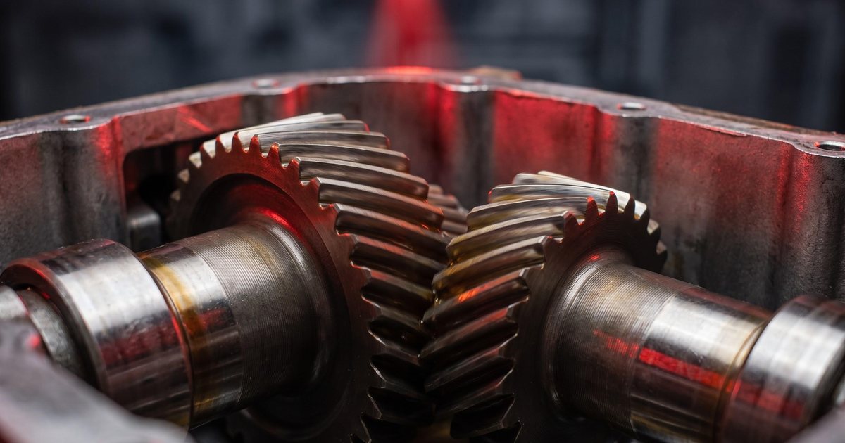 Two helical gears meshing inside a gearbox, coated in golden gear oil